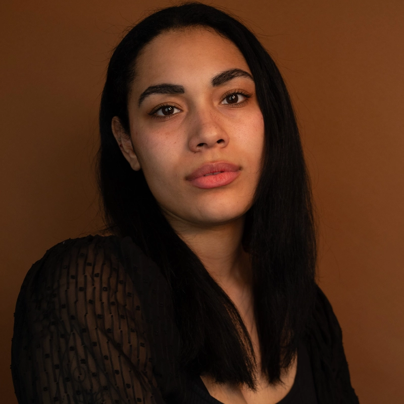 A woman with straight, long black hair and natural makeup gazes at the camera. She wears a black sheer dress with a dotted texture. The background is a solid warm brown, creating a simple and intimate portrait.
