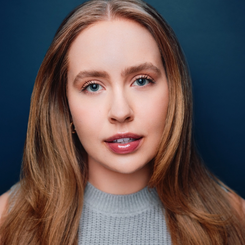 Isa Rodriguez, a young woman with long, light brown hair and blue eyes, looks directly at the camera. She wears a gray sleeveless knit top and natural makeup. The deep blue background gives the portrait a polished, professional feel.
