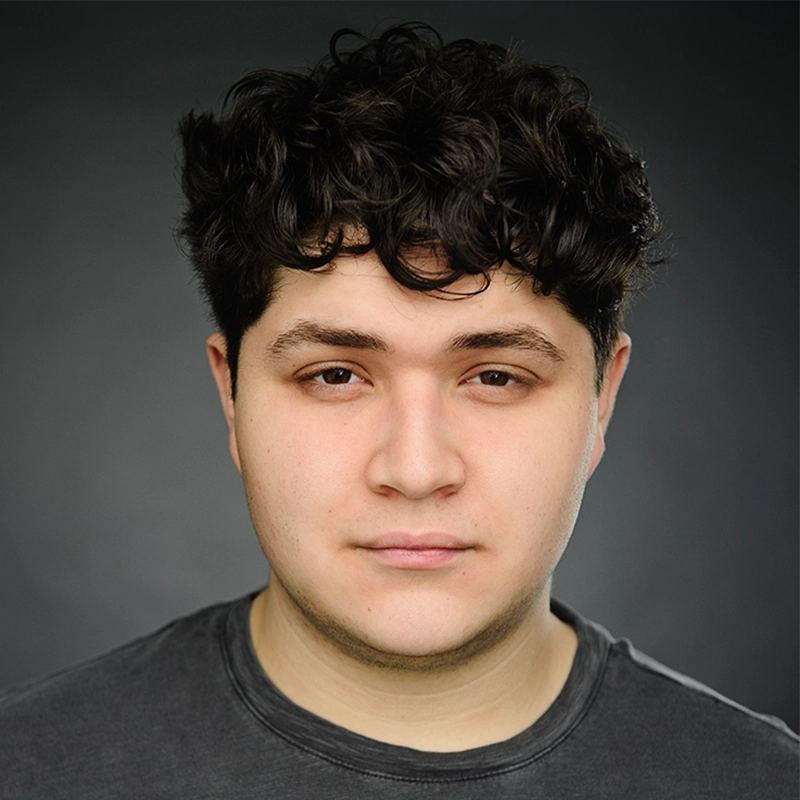 A young person with short, curly dark hair and fair skin faces the camera with a neutral expression. They wear a dark t-shirt and are set against a plain, softly lit dark background.