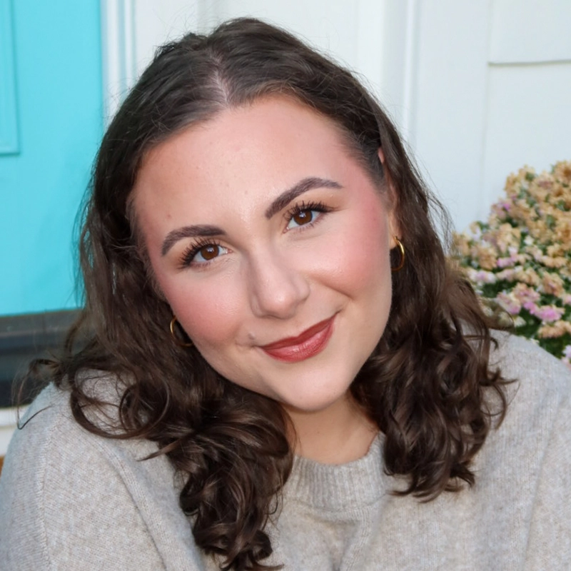 A woman with curly brown hair and gold hoop earrings smiles at the camera. She wears a beige sweater and natural makeup with rosy cheeks and brown lipstick. There’s a light blue door and a flowering plant in the background.