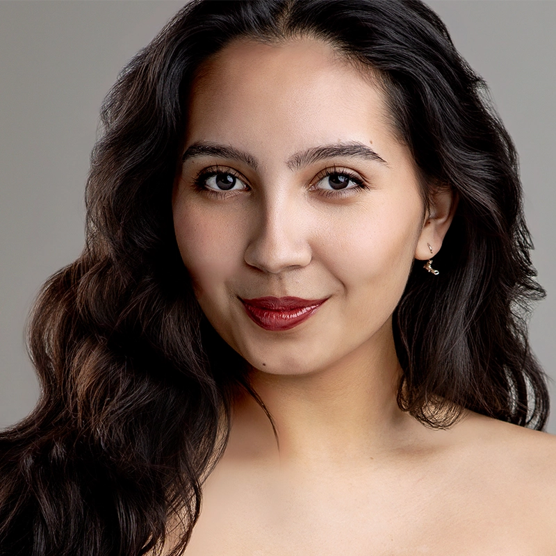 A young woman with long, wavy dark hair poses in front of a neutral gray background. She has medium skin tone, dark eyes, and wears subtle makeup with red lipstick. She is smiling softly and wearing small earrings, with bare shoulders visible.