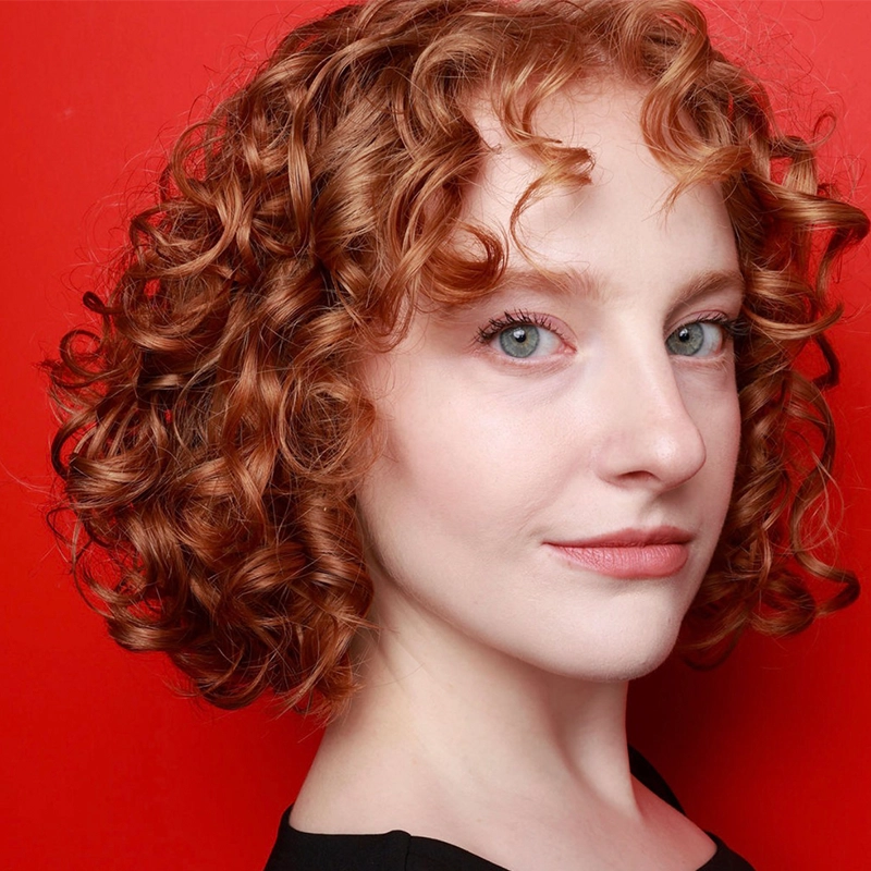 A woman with fair skin and short, curly red hair poses against a solid red background, wearing a black top. She has blue eyes, natural makeup, and a subtle smile, with her head slightly turned to the side, highlighting her curls.