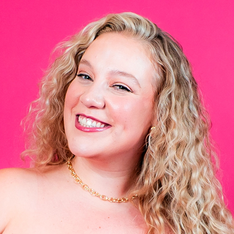 A smiling woman with long, curly blonde hair and fair skin poses against a bright pink background. She wears a gold chain necklace and hoop earrings. Her makeup is subtle, featuring glossy pink lipstick. She exudes a cheerful and confident demeanor.