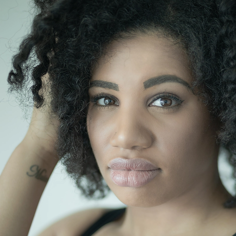 A woman with curly dark hair and defined eyebrows looks directly at the camera. She has full lips, smooth skin, and a soft expression. Tara Jackson touches her hair, revealing a tattoo on her inner forearm against a light, blurred background.