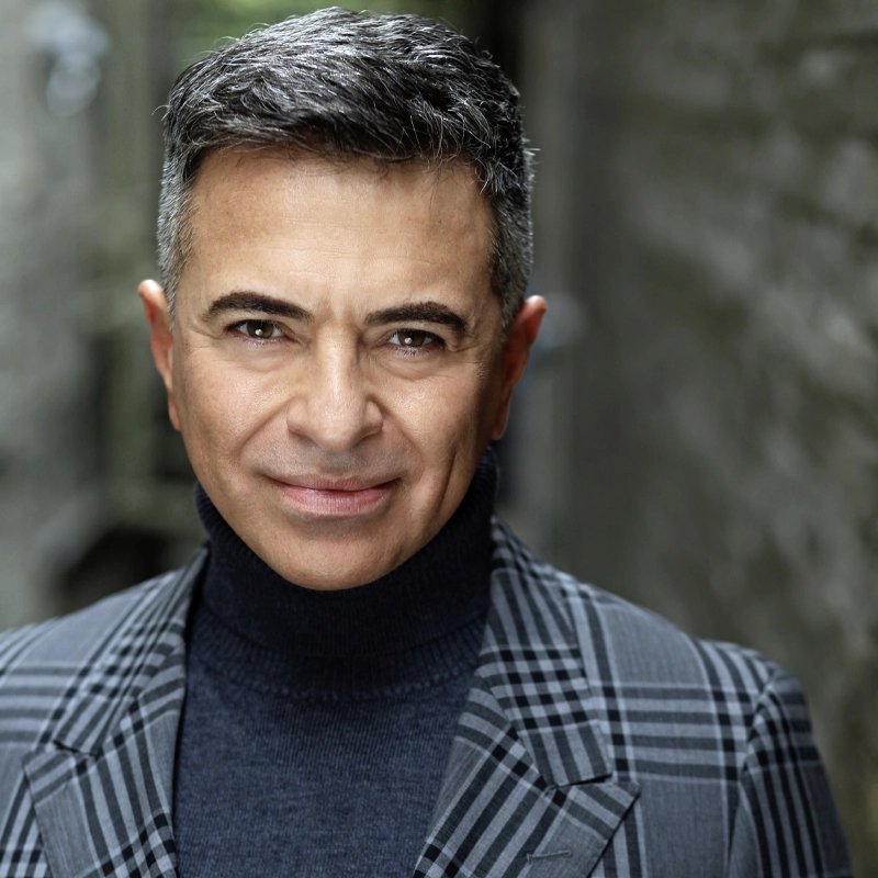 Luis Villabon, a middle-aged man with short, gray hair, smiles gently at the camera. He wears a dark turtleneck under a gray plaid suit jacket and stands in an outdoor, blurred alleyway with stone walls and soft lighting.
