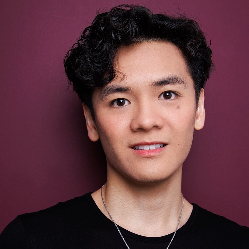 A young person with short, wavy black hair and smooth skin stands in front of a dark maroon background. They wear a black shirt, a silver necklace, and are smiling softly at the camera. The lighting highlights their facial features evenly.
