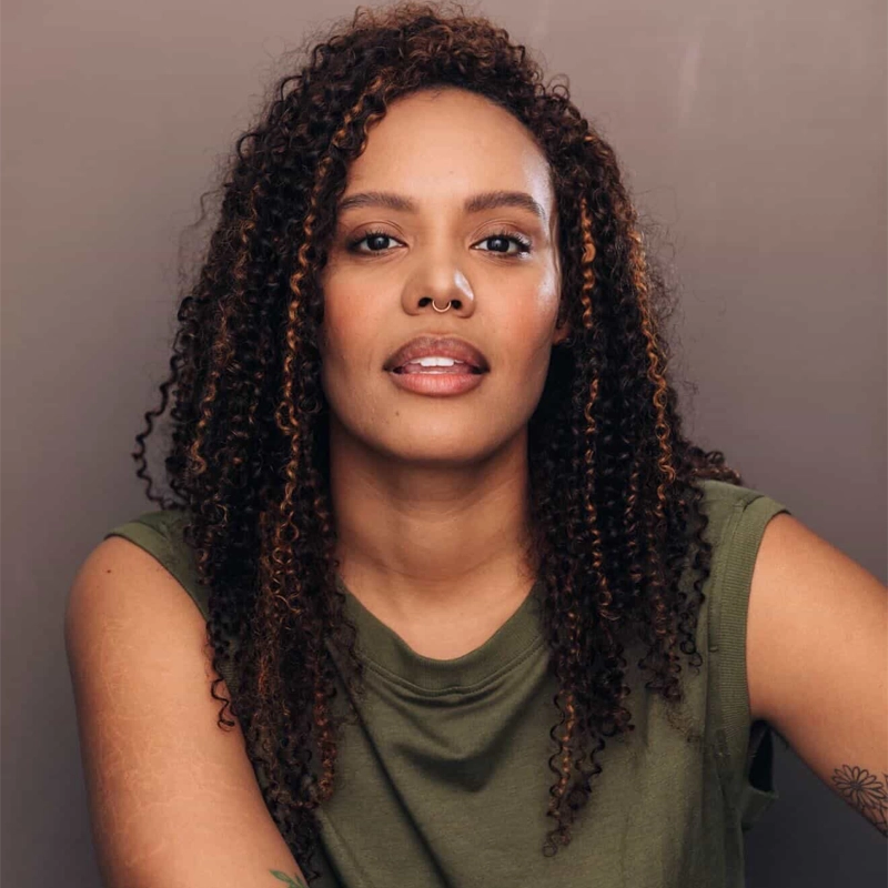 Miki Abraham, with long, curly hair and light brown skin, sits against a plain beige background. She wears a sleeveless olive green top and looks confidently into the camera, her relaxed expression complemented by partially visible arms in the foreground.