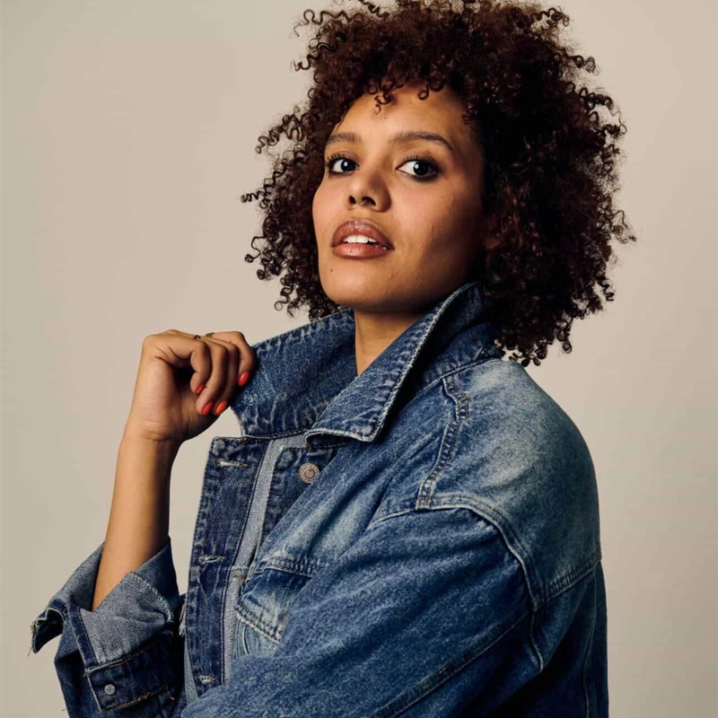 Miki Abraham, with curly hair and medium skin tone, looks confidently at the camera. Wearing a large denim jacket with the collar raised, they hold it with one hand against a plain background, exuding calm and self-assured energy.