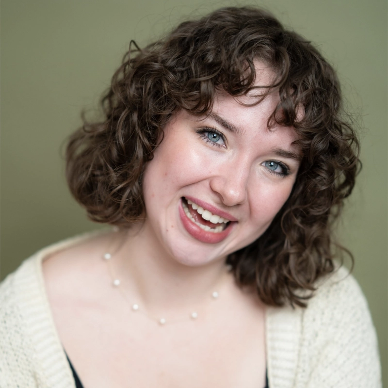 A young woman with short, curly brown hair smiles at the camera. She has blue eyes, light skin, and is wearing a cream knit sweater and a delicate pearl necklace. The background is a muted green. Her expression is cheerful and friendly.