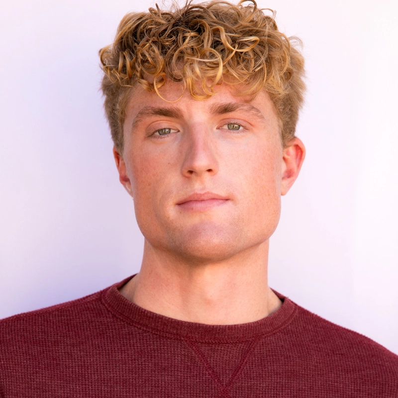 Nolan Montgomery, a young man with curly blond hair and green eyes, looks at the camera with a neutral expression. He is wearing a maroon crew-neck sweater against a plain, light-colored background, giving the image a clean, simple feel.
