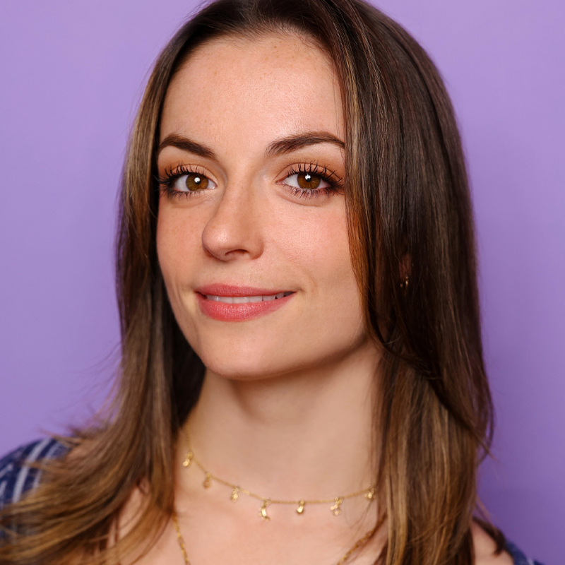 Claire-Frances Sullivan, a young woman with straight brown hair and brown eyes, smiles gently at the camera. She wears a gold necklace with small charms and a blue top. The background is light purple; her makeup is natural and expression calm.