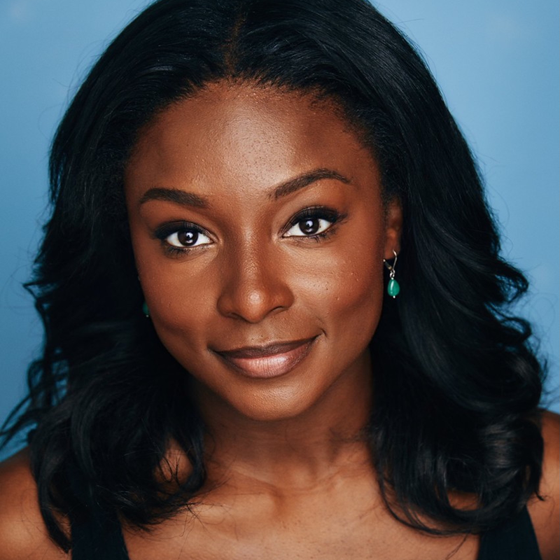 Taylor-Marie Daniel, a woman with dark wavy hair and brown skin, smiles softly against a bright blue background. She wears green earrings and a black top, her natural makeup enhancing her clear skin and expressive brown eyes.