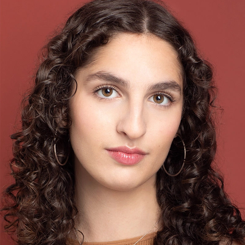 Aidan S. Wells, a young person with long, dark, curly hair and hoop earrings, poses calmly against a reddish background. With light skin and full lips in natural pink gloss, they wear a neutral top and gaze confidently at the camera.