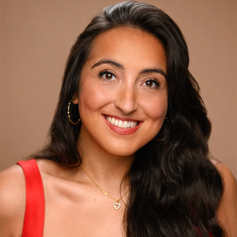 Shelby Acosta, with long, wavy dark hair, smiles at the camera in a sleeveless red top, gold hoop earrings, and a gold heart-shaped necklace. The warm brown background highlights her natural makeup and rosy lips.