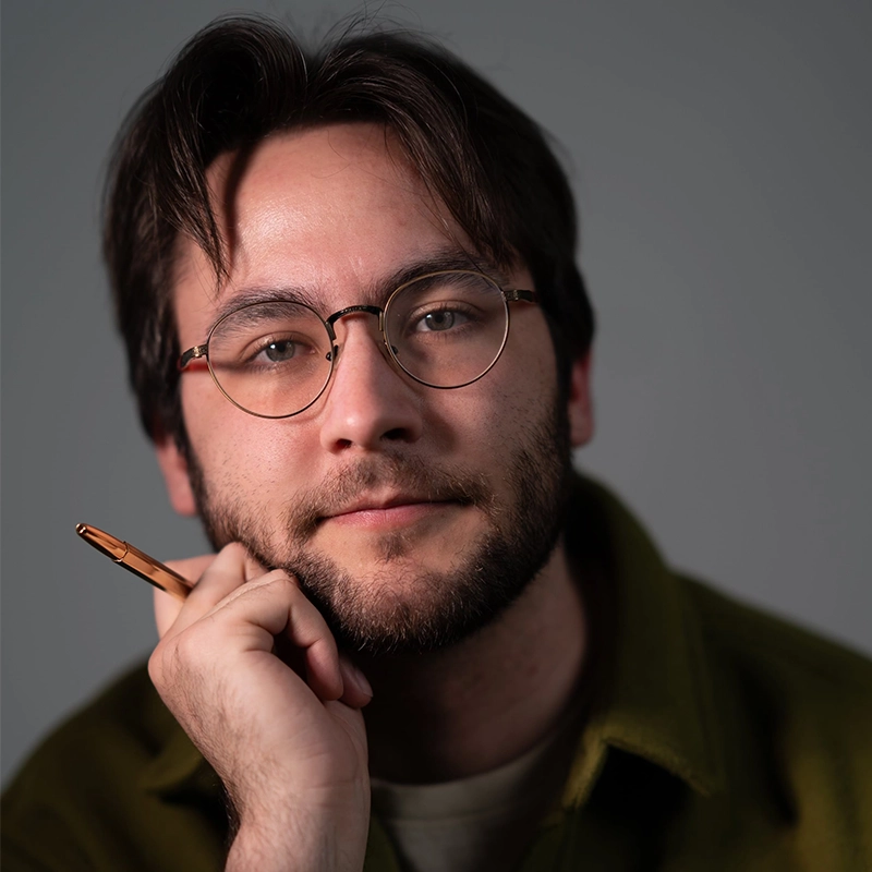 Aidan S. Wells, a young man with brown hair, glasses, and a beard, looks thoughtfully at the camera. He wears a green jacket over a beige shirt and holds a pen to his chin against a plain gray background with soft, even lighting on his face.
