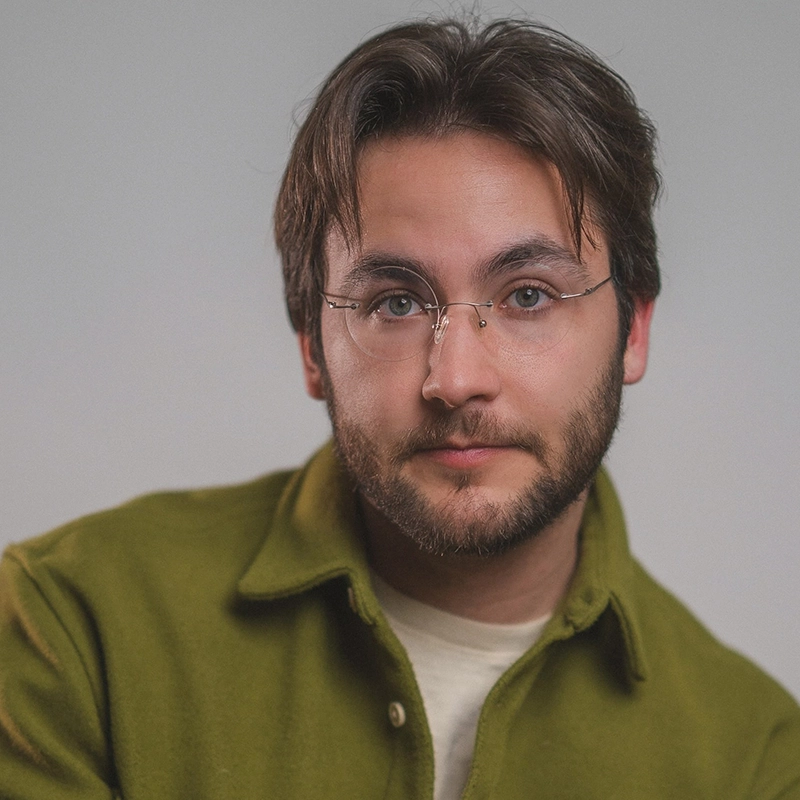 Aidan S. Wells, a young man with short brown hair, a trimmed beard, and glasses, looks at the camera with a slight smile. He wears a light green jacket over a cream-colored shirt against a plain, softly lit background that emphasizes his face.
