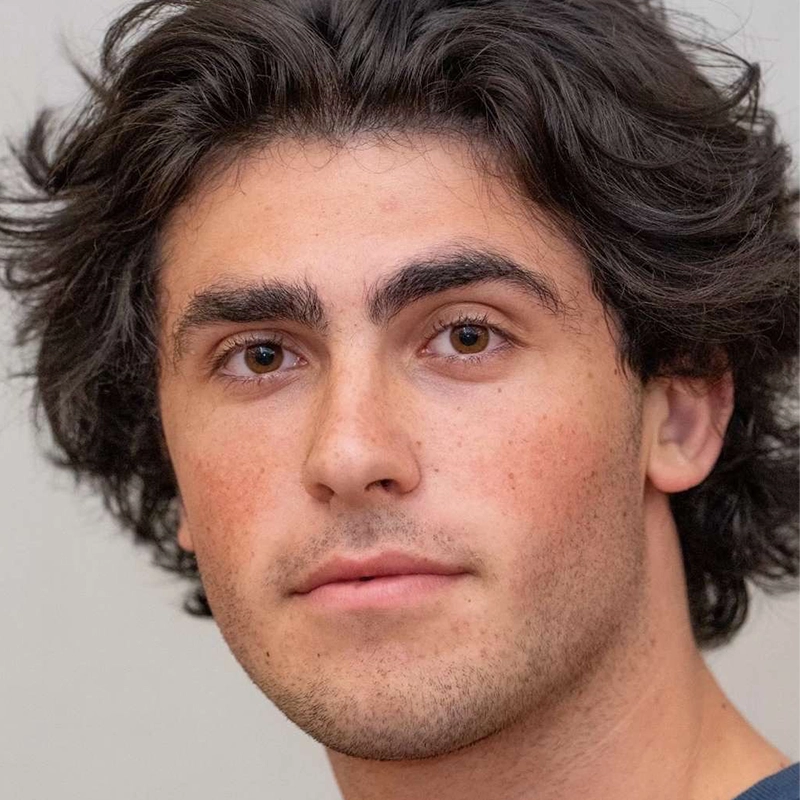 Aidan S. Wells, a young man with wavy dark hair and thick eyebrows, looks directly at the camera with a neutral expression. He has light skin and brown eyes, set against a plain, light-colored background.