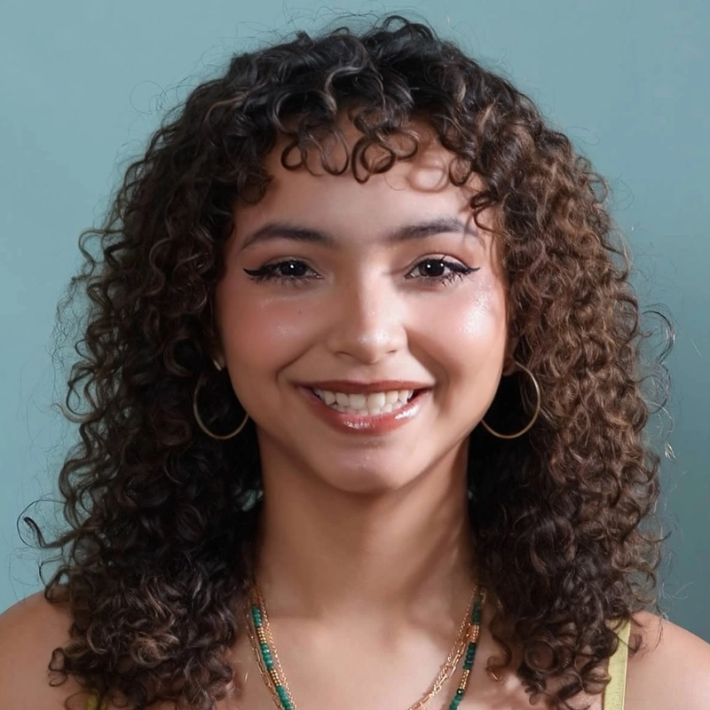 Aidan S. Wells captures a young woman with curly brown hair, gold hoop earrings, and layered necklaces, smiling at the camera. She sports winged eyeliner, natural makeup, a light green top, and stands against a solid teal background.