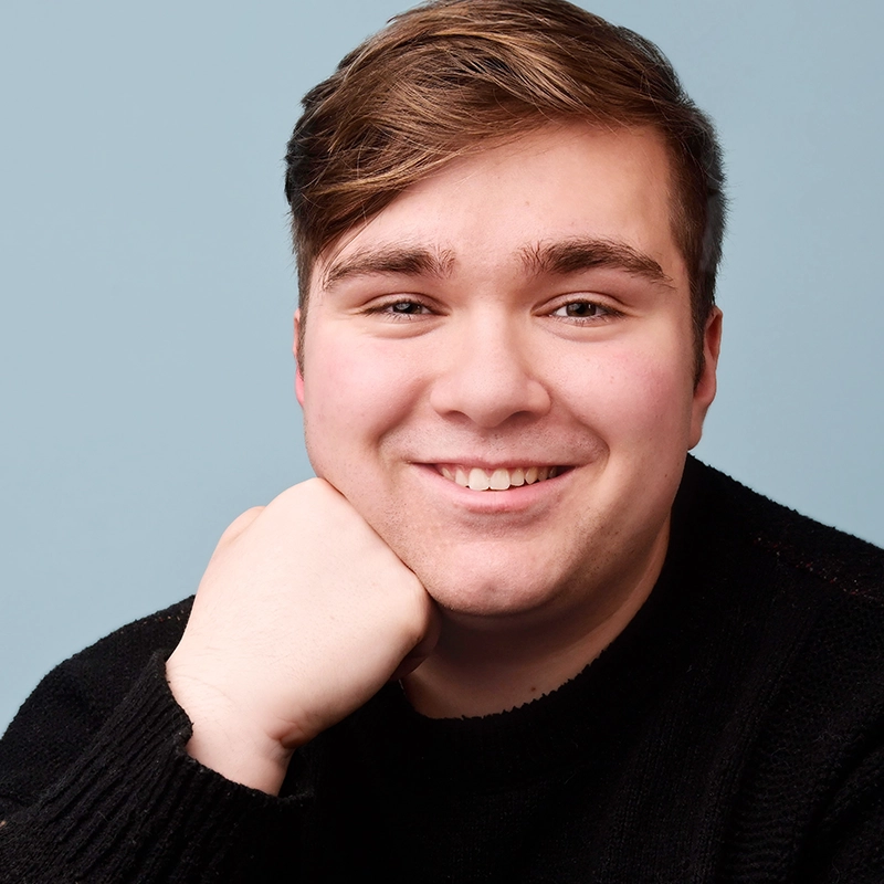 Aidan S. Wells, a young man with light skin and short brown hair, smiles warmly at the camera. He is wearing a black sweater and resting his chin on his hand against a solid light blue background, creating a calm and friendly feel.
