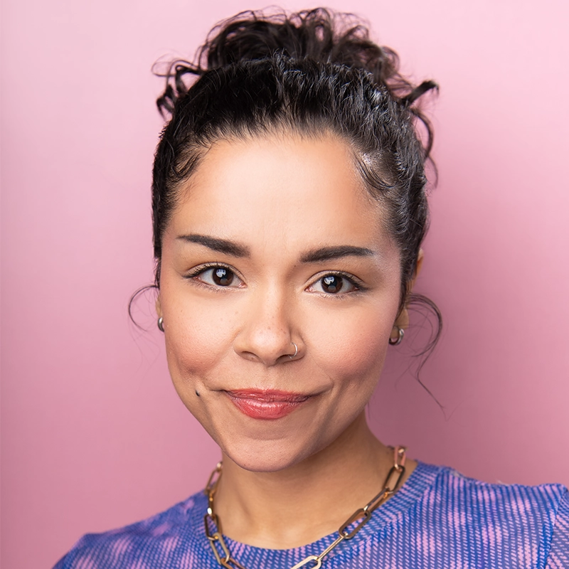 Brooke Simpson, with medium skin tone, dark curly hair in a bun, and a nose ring, smiles softly. She wears a purple top and chunky gold chain necklace. The solid light pink background gives the portrait a bright and cheerful feel.