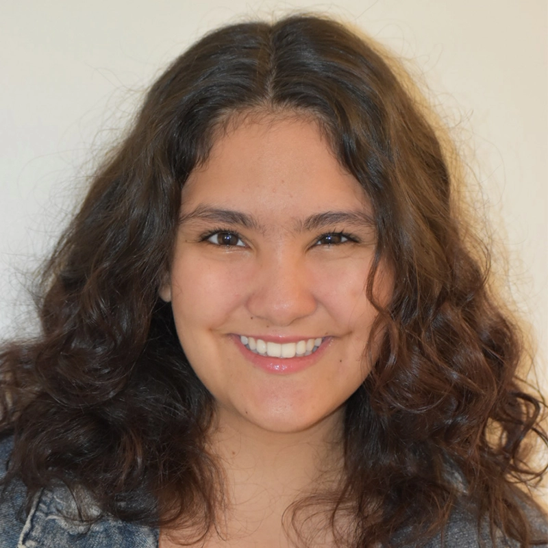 Aidan S. Wells captures a young woman with wavy brown hair and medium skin smiling warmly at the camera. She has dark eyes and wears a denim jacket over a light top, set against a plain, light-colored background that highlights her face.