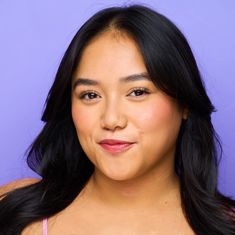 A young woman with long, dark hair and smooth skin smiles softly at the camera. With natural makeup and rosy cheeks, she wears a light purple top against a solid lavender background, in a style reminiscent of Aidan S. Wells' portraits.