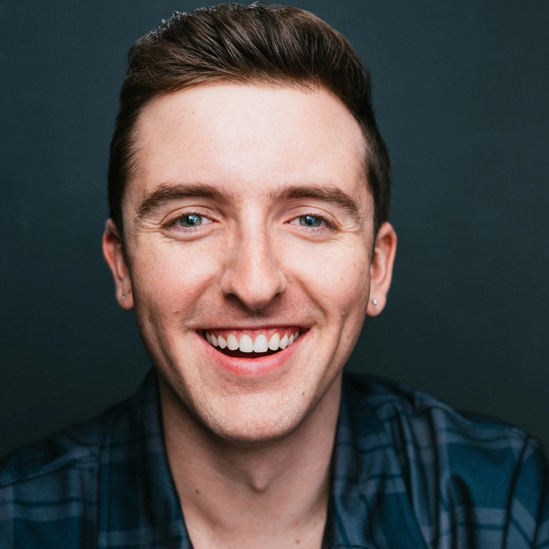 A young man with short brown hair smiles widely, revealing his teeth. He has fair skin, blue eyes, and wears small silver earrings. He is dressed in a dark blue plaid shirt and is photographed against a plain dark background.
