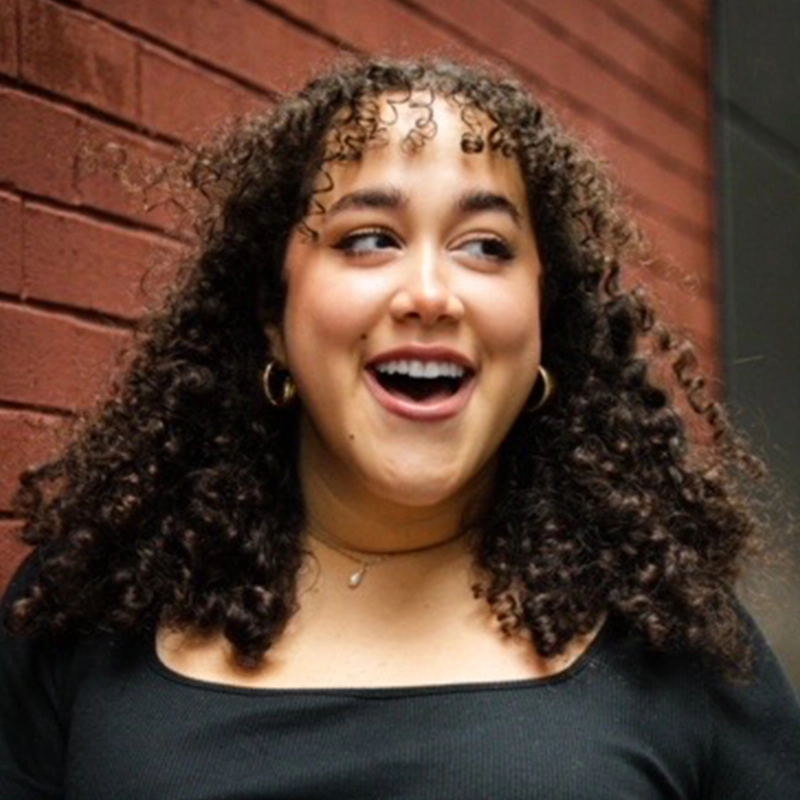 Aidan S. Wells captures a young woman with curly brown hair and gold hoop earrings smiling widely, looking to her left. She wears a black top and a delicate necklace, with a brick wall and part of a gray structure behind her.