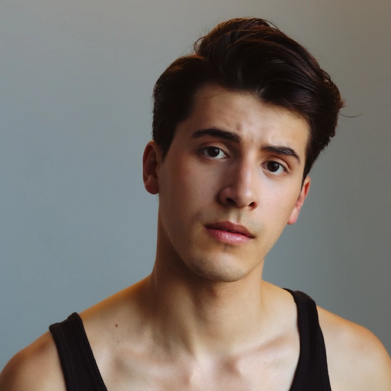 Aidan S. Wells, a young man with short, dark brown hair and fair skin, wears a black tank top. He gazes slightly to the side with a neutral, thoughtful expression against a softly lit, light gray background.