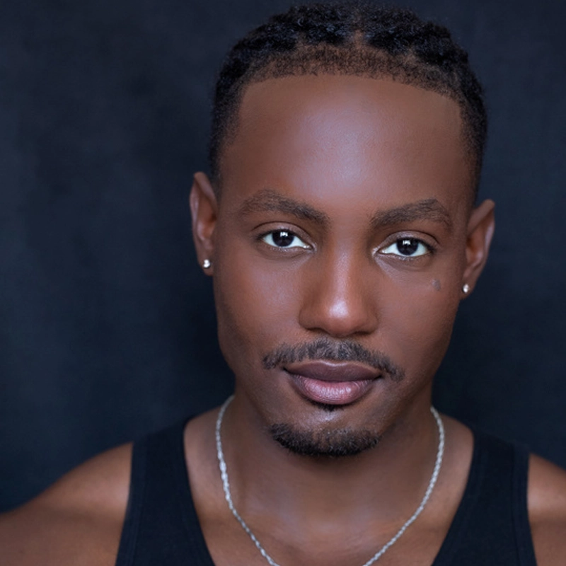 A man with short, braided hair and trimmed facial hair looks directly at the camera. He wears a black tank top, silver necklace, and small stud earrings. His expression is neutral and the background is dark.