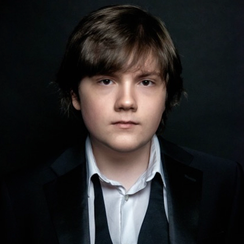 A young person with straight brown hair wears a black suit jacket, untied black bowtie, and white shirt. They face forward with a neutral expression. The background is dark, highlighting Ethan Mathias’s face and formal attire.