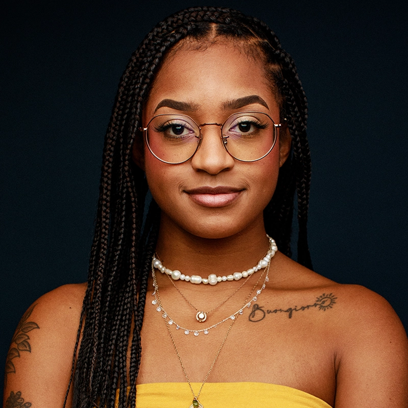 Olivia Hardy, a young woman with long braids, round glasses, and layered necklaces, poses confidently against a dark background. She wears a yellow top, sun tattoo and script on her shoulder, and offers a slight smile to the camera.
