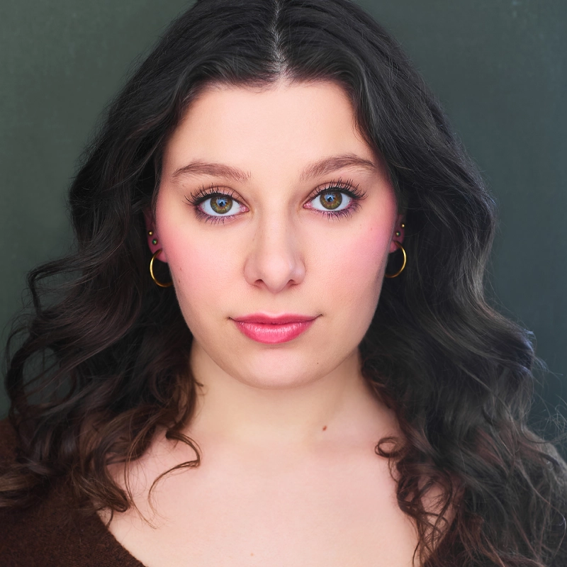 A young woman with wavy dark hair and fair skin, Lauren Riddle wears gold hoop earrings and looks directly at the camera. She has blue-green eyes, natural makeup, and a slight smile, posed against a dark, softly blurred background.