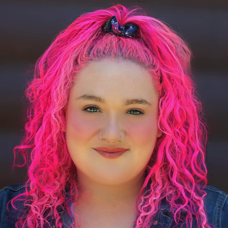 A person with bright pink, curly hair tied up with a scrunchie. They have light skin, wear subtle makeup with pink blush, and have two nose piercings. They are dressed in a denim jacket and are smiling softly against a dark background.