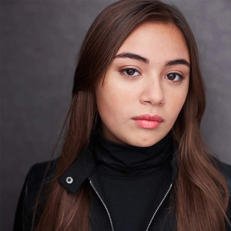 Scarlett Maikish, a young woman with long brown hair, straight eyebrows, and light skin, gazes seriously at the camera. She wears a black turtleneck and jacket, standing against a plain, softly textured gray background.