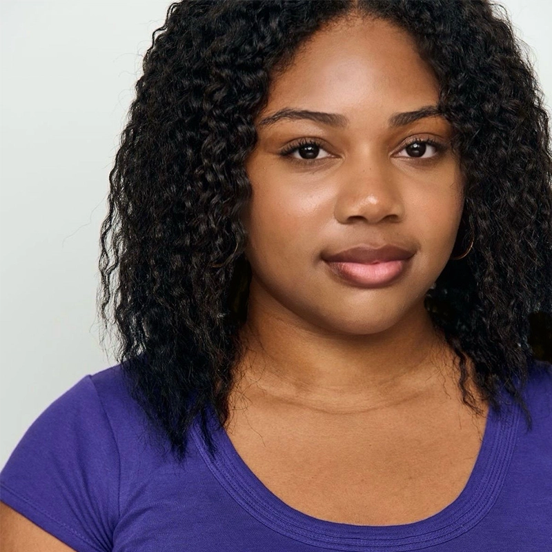 A young woman with medium skin tone and curly, shoulder-length black hair faces the camera. She is wearing a purple top and small hoop earrings. The background is plain and light-colored, creating a simple, clean portrait.