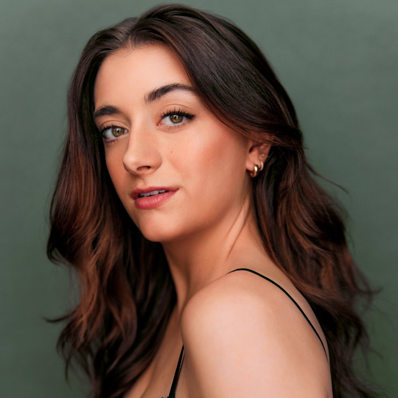 Natalie Steele, a woman with wavy brown hair and light skin, poses in front of a muted green background, looking over her shoulder. She wears a black spaghetti strap top, subtle makeup, and small gold hoop earrings, exuding confidence and poise.