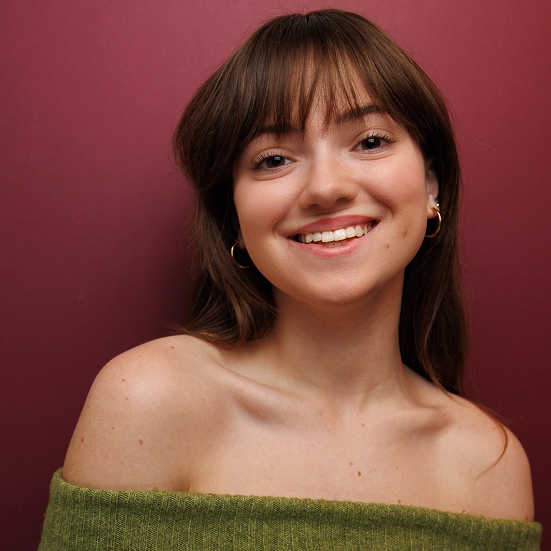 A young woman with long brown hair and bangs smiles warmly at the camera. She wears gold hoop earrings and an off-shoulder green top. The solid burgundy background adds to the inviting mood of this Aidan S. Wells inspired portrait.