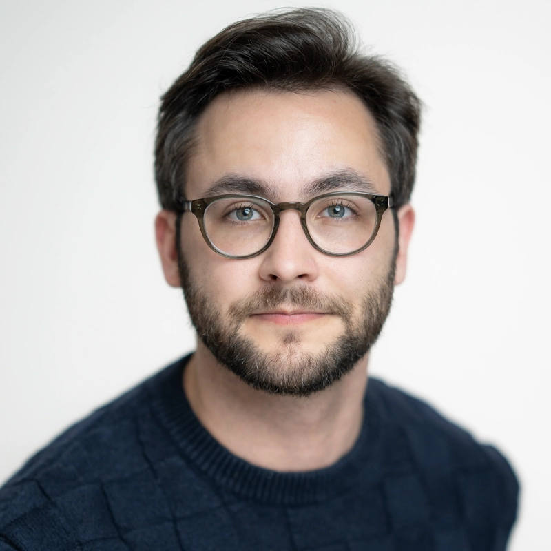 A portrait of Aidan S. Wells, a man with a beard, dark hair, and light-colored eyes wearing clear-framed glasses. He is dressed in a dark blue, textured knit sweater against a plain white background.