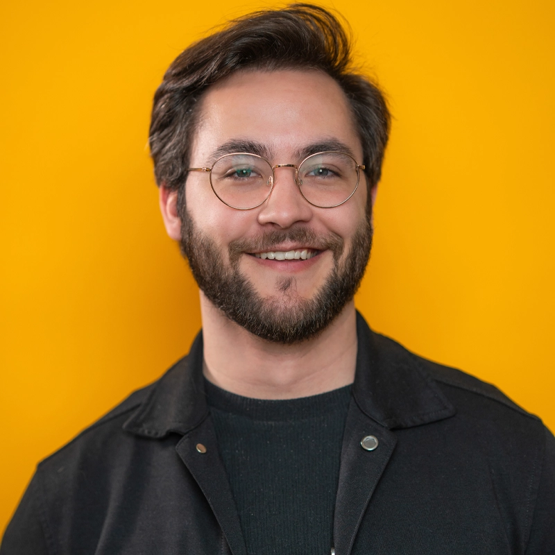 Aidan S. Wells, a smiling man with dark hair, beard, and glasses, stands against a bright yellow background. He wears a black jacket over a dark shirt, and his expression is friendly and approachable.