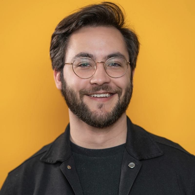 Aidan S. Wells, a man with short brown hair, a beard, and glasses, smiles at the camera. He is wearing a black jacket over a dark shirt. The background is solid bright yellow.
