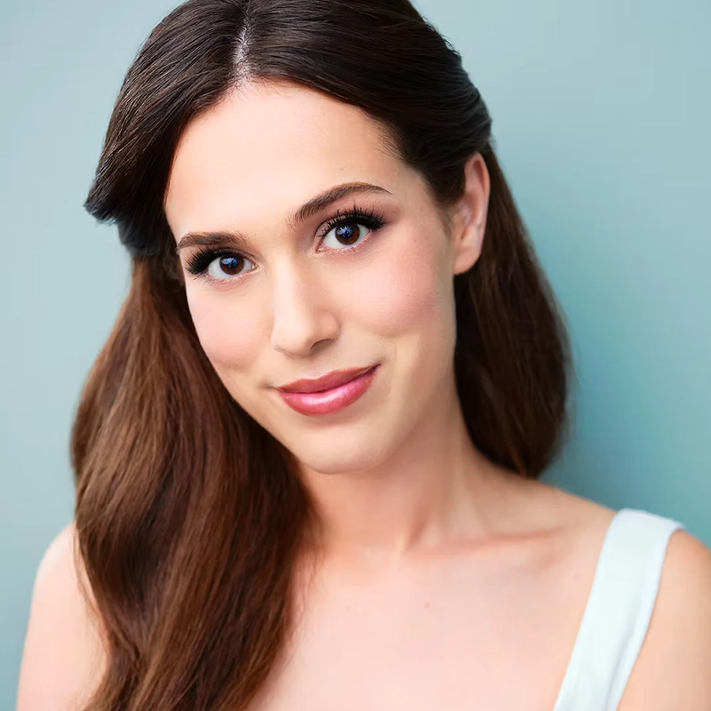 Alyssa Jaffe, a young woman with long, brown, wavy hair styled back, smiles softly at the camera in a sleeveless white top. She has clear skin, natural makeup with defined eyelashes, and is posed against a soft teal background.