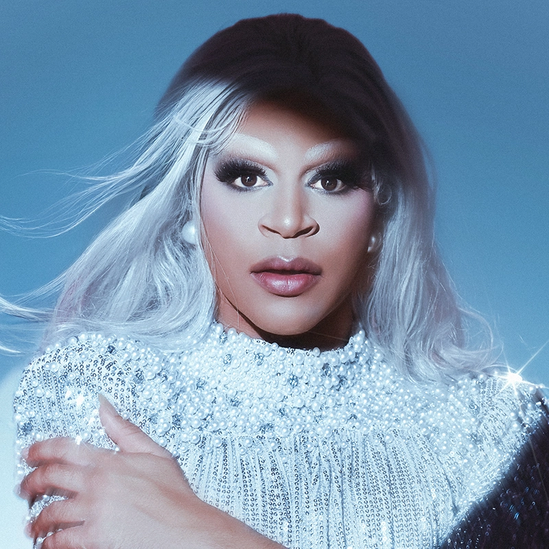 Alyssa Jaffe, with long, wavy silver hair and dramatic makeup, wears a sparkling sequined top. Pearl earrings accent the look as she poses confidently against a soft blue background, her hair wind-swept and expression self-assured.