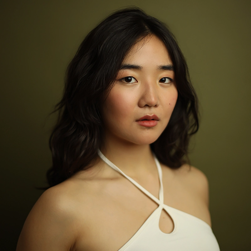 A young woman with long, wavy dark hair poses against an olive green background. She wears a cream halter top with a keyhole design, has subtle makeup, and a calm, neutral expression while looking slightly off-camera.
