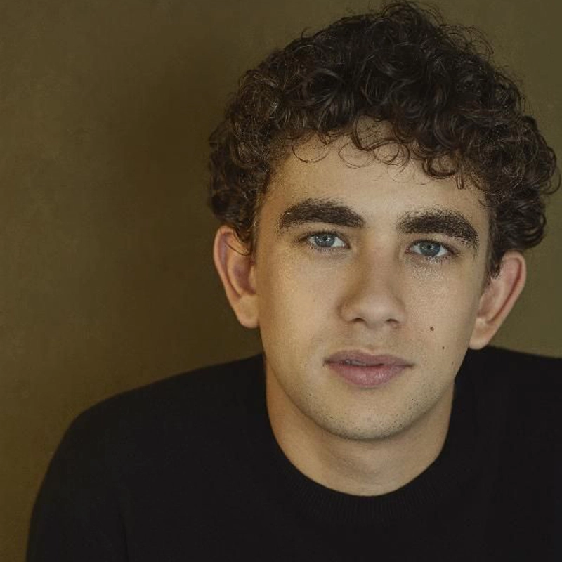 A portrait of a young person with curly dark hair and blue eyes, wearing a black shirt against a plain background.