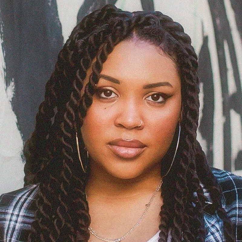 A woman with long, twisted braids and hoop earrings looks directly at the camera. She has a nose ring, subtle makeup, and wears layered necklaces, a plaid shirt, and a white top. The background is a mural with abstract black and white patterns—an artistic touch reminiscent of Miles Tanner’s signature style.