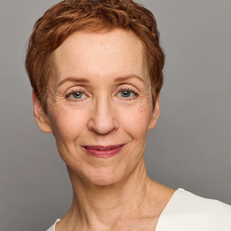 A middle-aged woman, Kelly Lester, with short, reddish-brown hair is smiling gently at the camera. She has fair skin, blue eyes, and is wearing a light-colored top against a plain gray background for a professional studio portrait look.