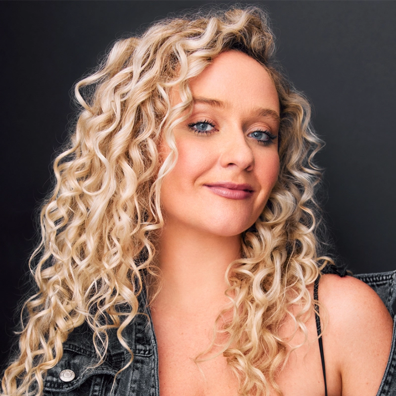 Alyssa Jaffe, with long, blonde, tight curly hair, poses confidently against a dark background. She has light skin and blue eyes, wearing a sleeveless black top and denim jacket draped over her shoulder, smiling softly with a relaxed expression.