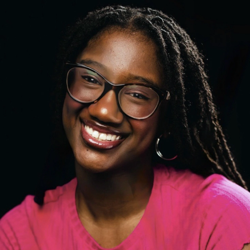 A smiling woman, Tsehai Marson, with dark skin, long black braided hair, and glasses wears a bright pink top. She has hoop earrings and stands against a plain black background, creating a vivid contrast with her clothing and warm smile.
