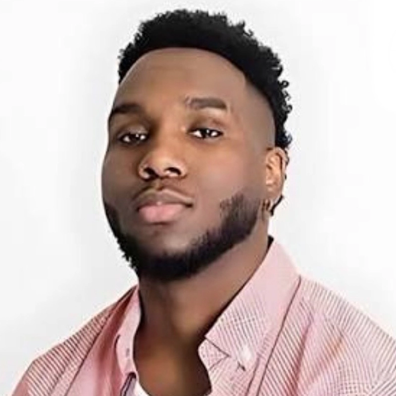 A young man, Miles Tanner, with short, curly black hair and a trimmed beard looks at the camera. He wears a pink button-up shirt over a white T-shirt and a small hoop earring. The background is plain and white.